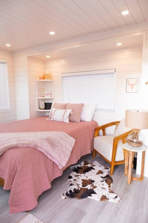 A bedroom with a bed, a chair, a lamp, a cowhide rug, and a shelf with items. - New! Cowgirl Shipping Container Home (Alpine)