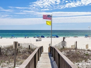 A wooden boardwalk leading to a beach with umbrellas and a flag. - Romar Towers 3B | Gulf-Front Condo Sleeps 6 (Orange Beach)