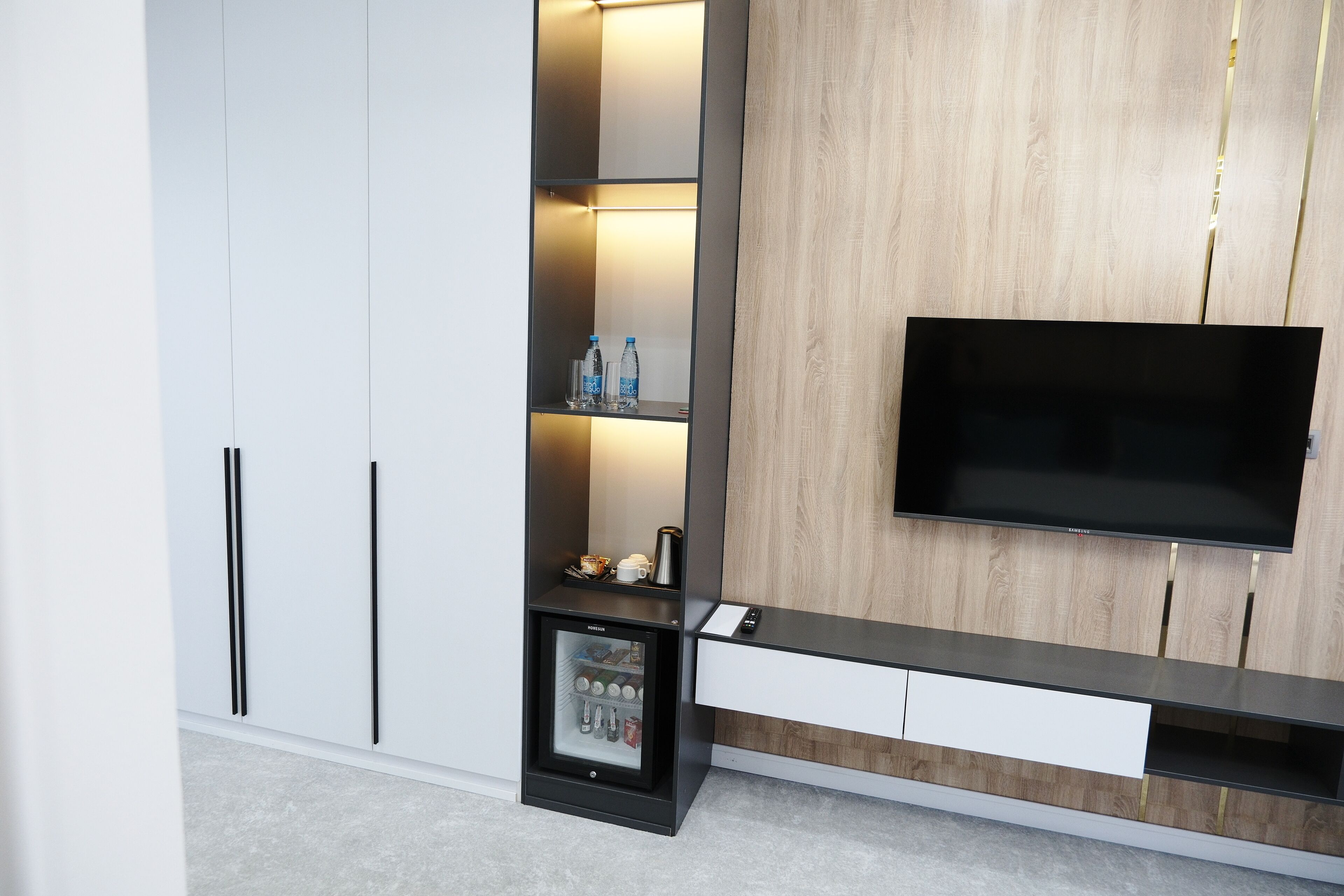 A modern living room with a built-in TV, a mini-fridge, and a shelf with bottled water and glasses.