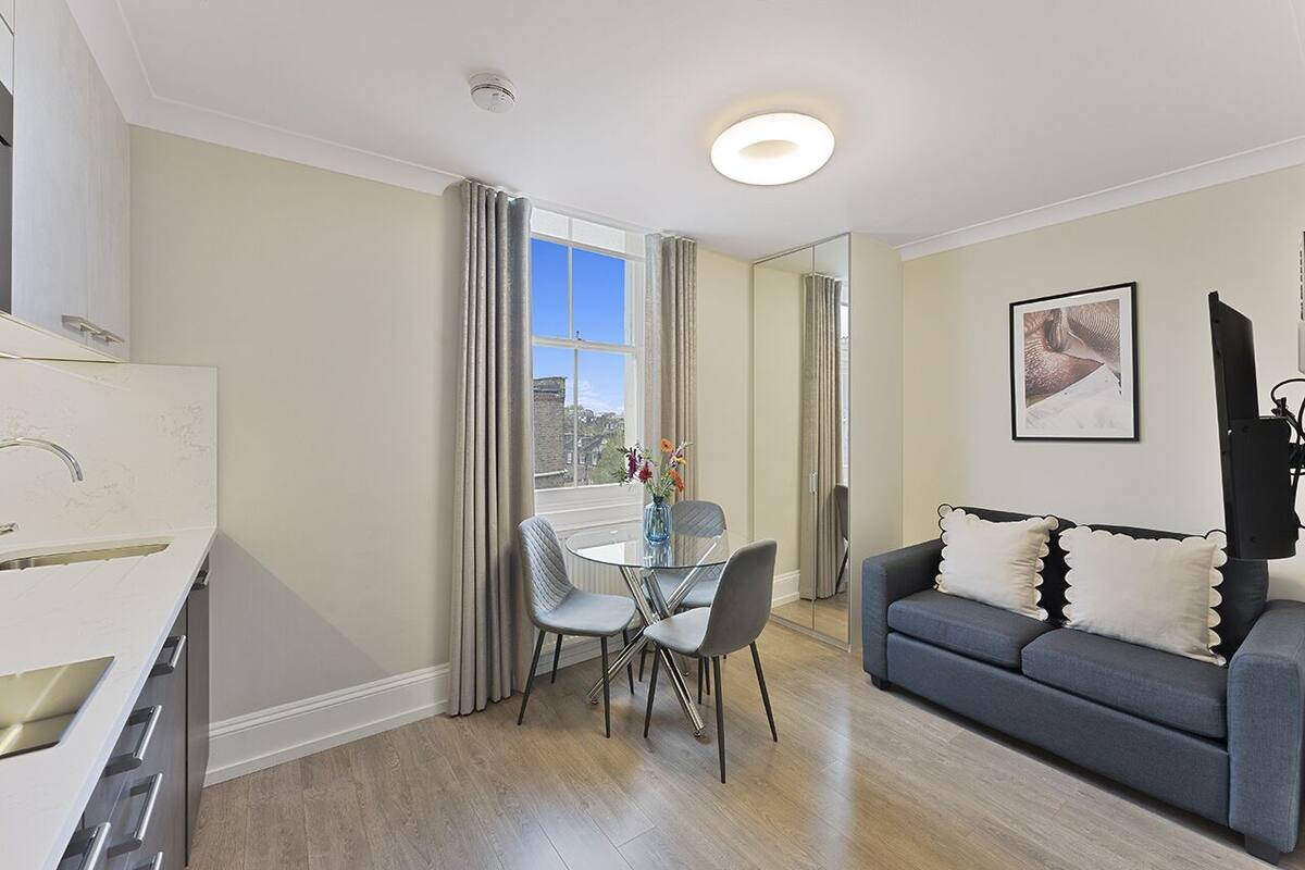 A modern living room with a grey sofa, a dining table with chairs, a kitchen area with a sink, and a large window with curtains.