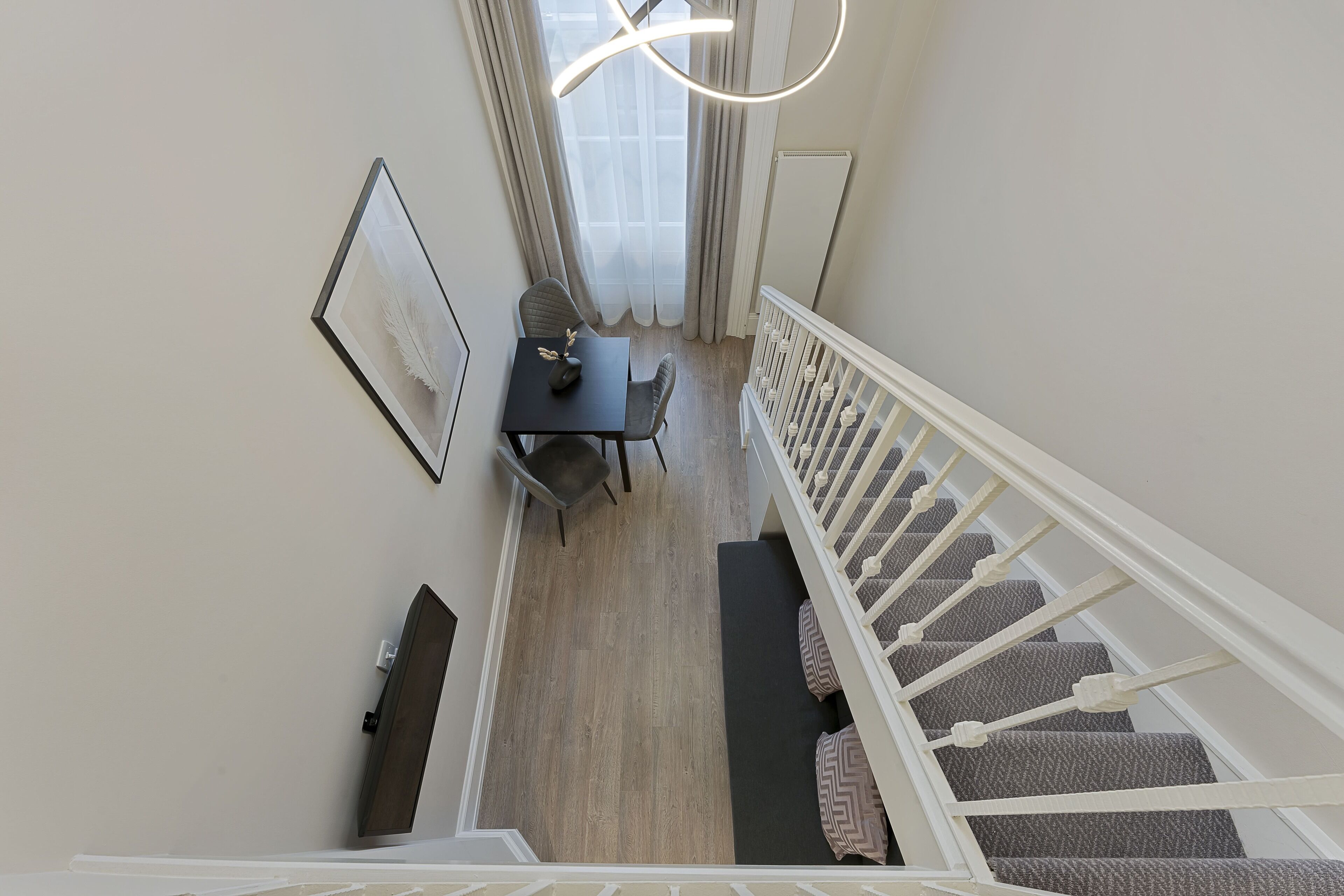 A staircase with a white railing leading to a landing. There is a black table with two chairs and a framed picture on the wall.