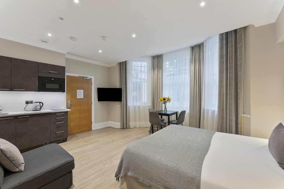 A modern hotel room with a bed, a sofa, a dining table with chairs, a TV, and a kitchen area.