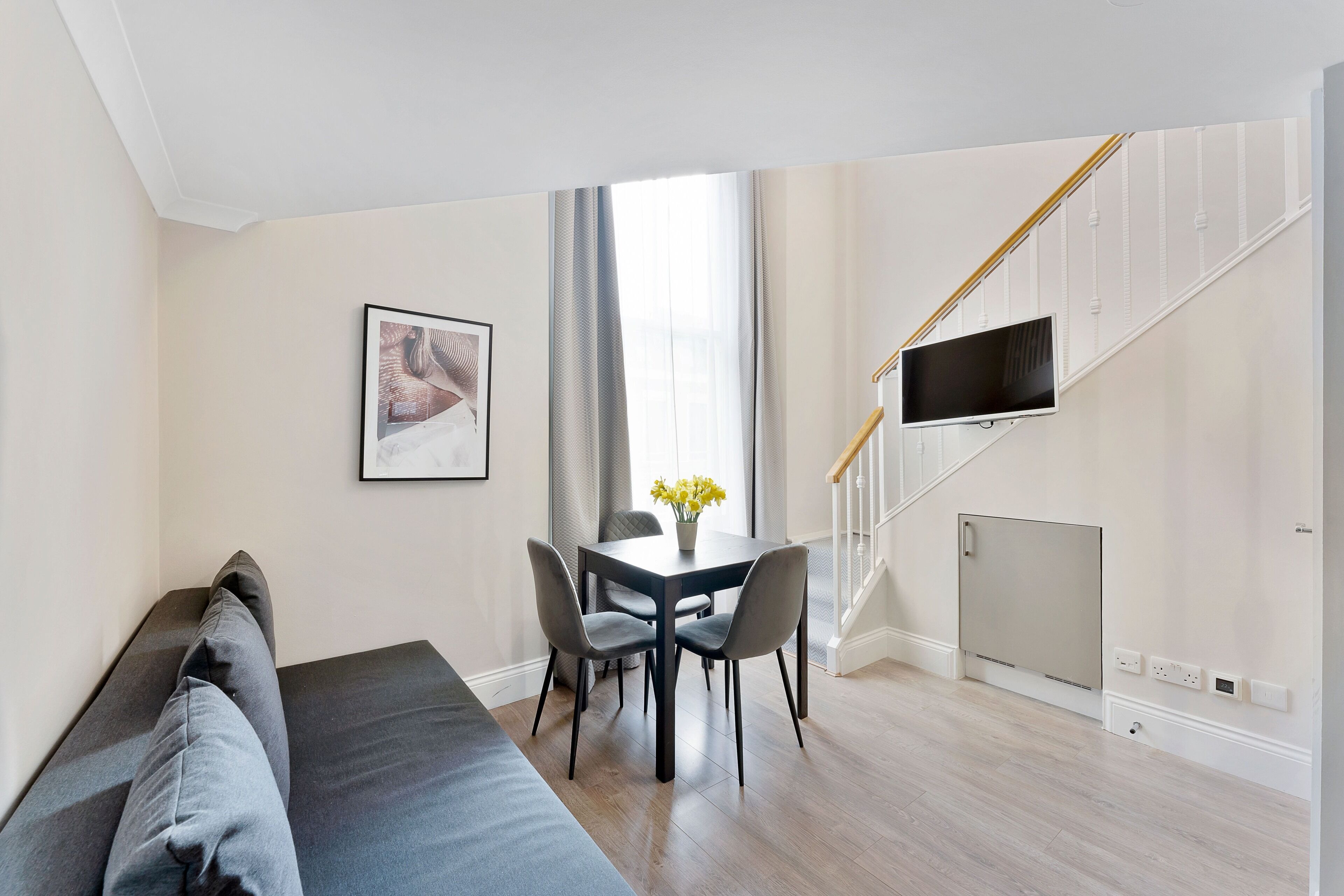 A modern living room with a sofa, dining table, and chairs. A TV is mounted on the wall, and there is a staircase with a railing.