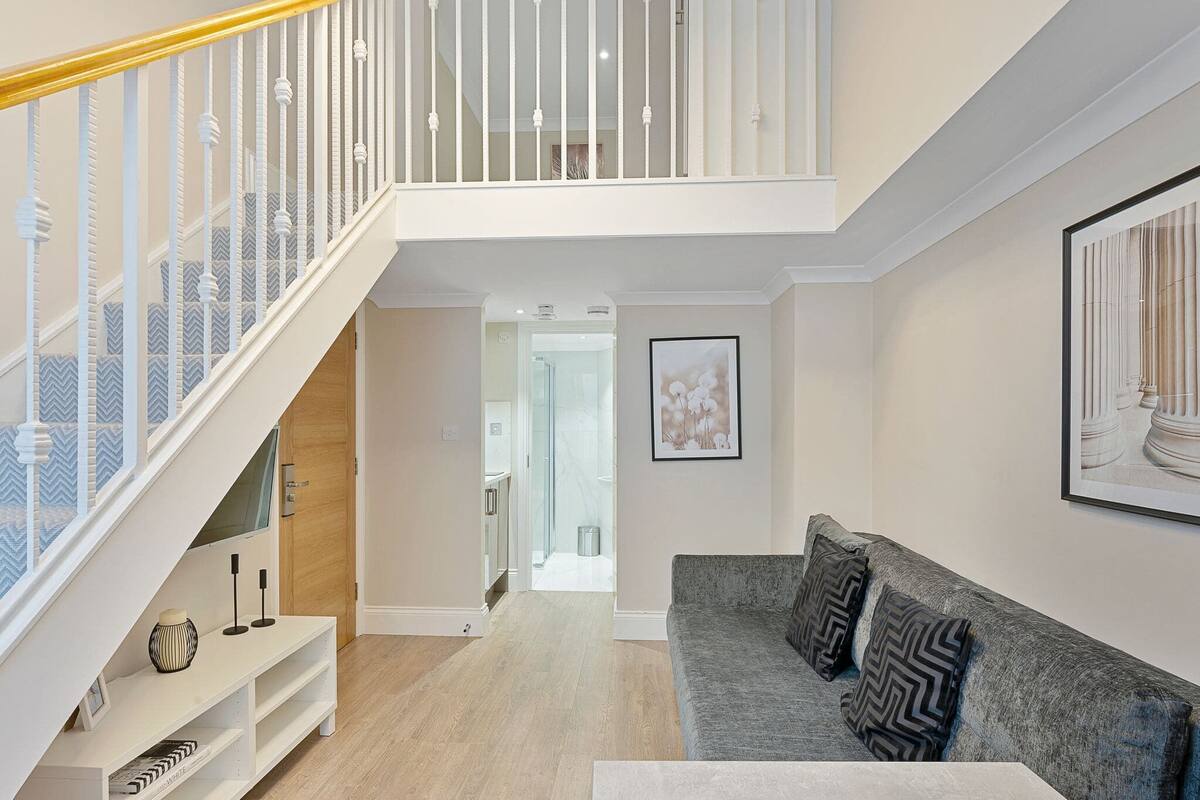 A modern living room with a grey sofa, a white entertainment unit, and a staircase with white railings.