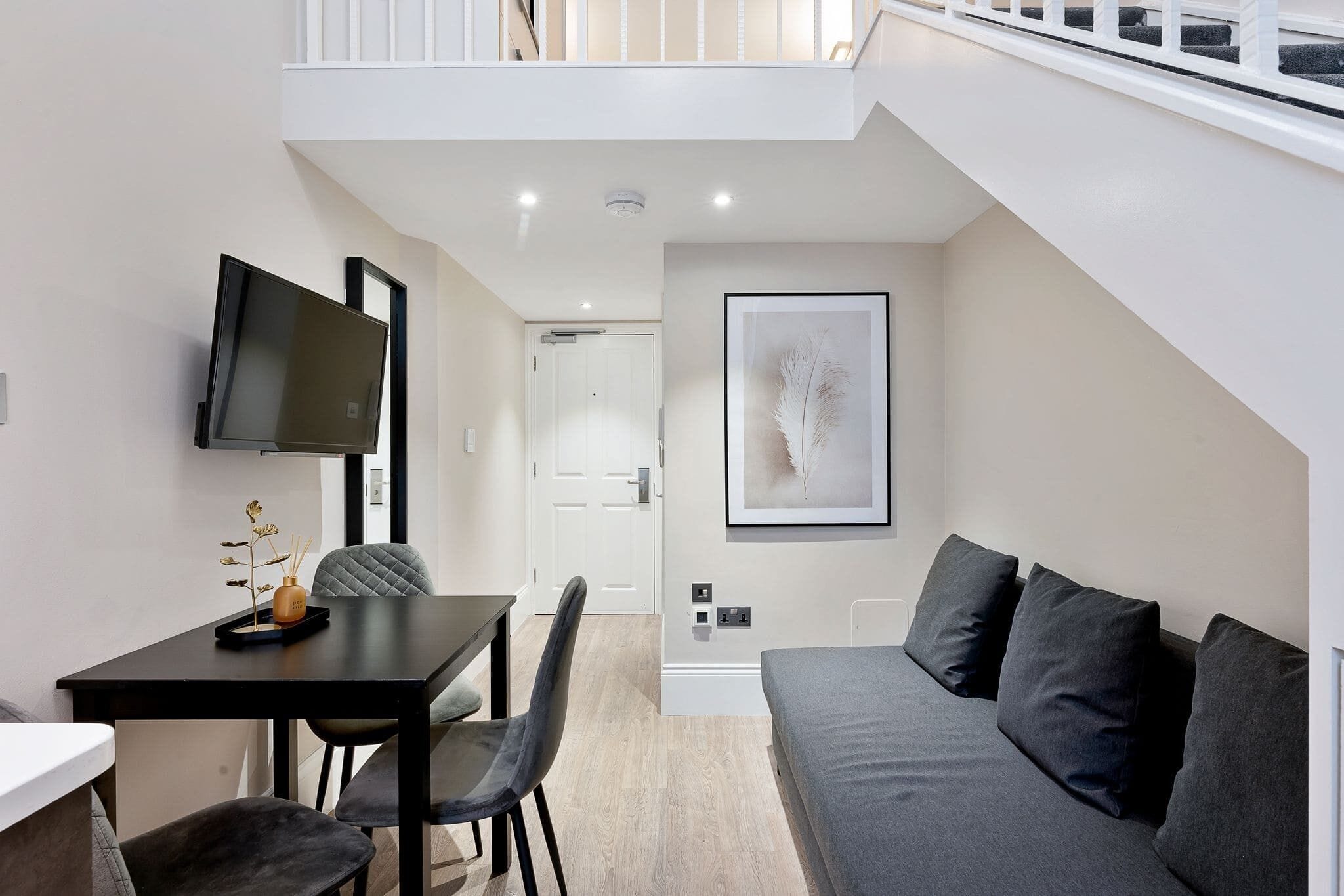 A modern living room with a sofa, a dining table with chairs, a flat-screen TV mounted on the wall, and a framed artwork above the sofa.