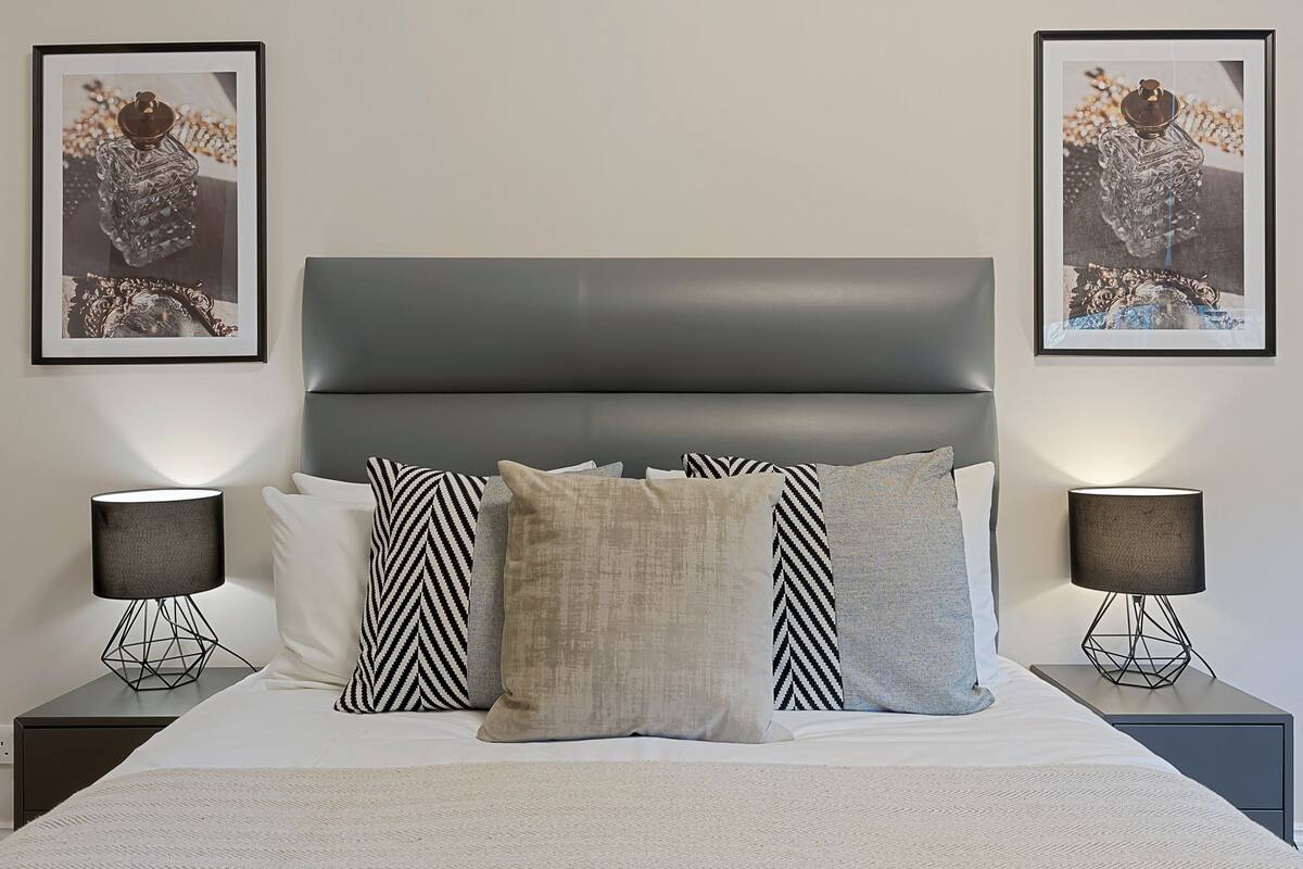 A neatly made bed with a grey headboard, flanked by two nightstands with lamps and framed pictures on the wall.