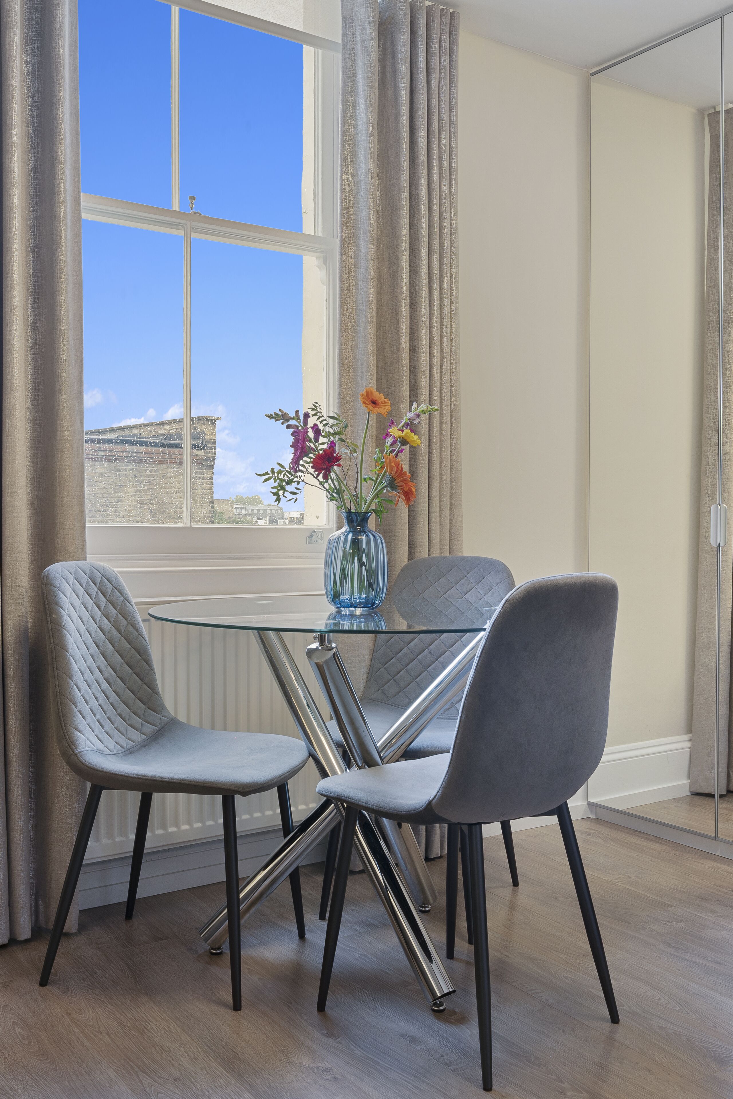 A dining area with a glass table and two chairs, a vase with flowers, and a window with curtains.