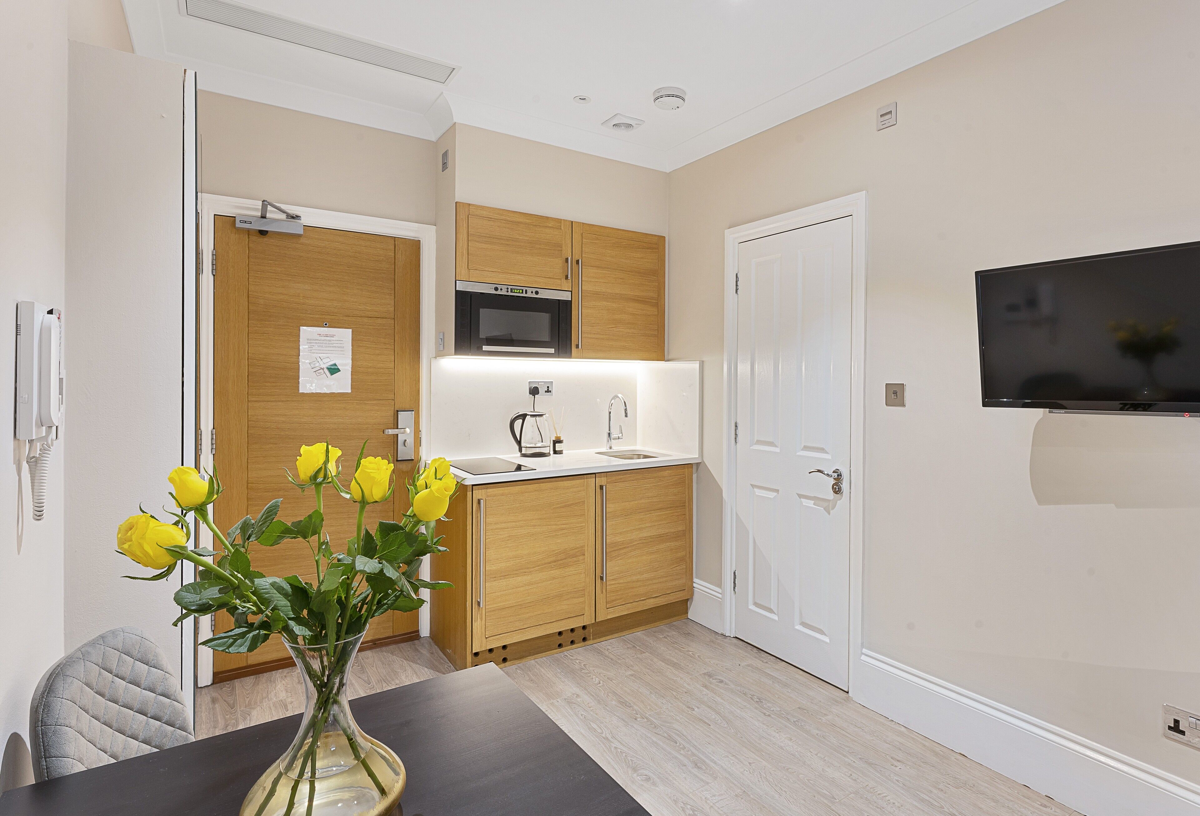A modern kitchen with wooden cabinets, a microwave, and a sink. There is a vase with yellow flowers on the table.