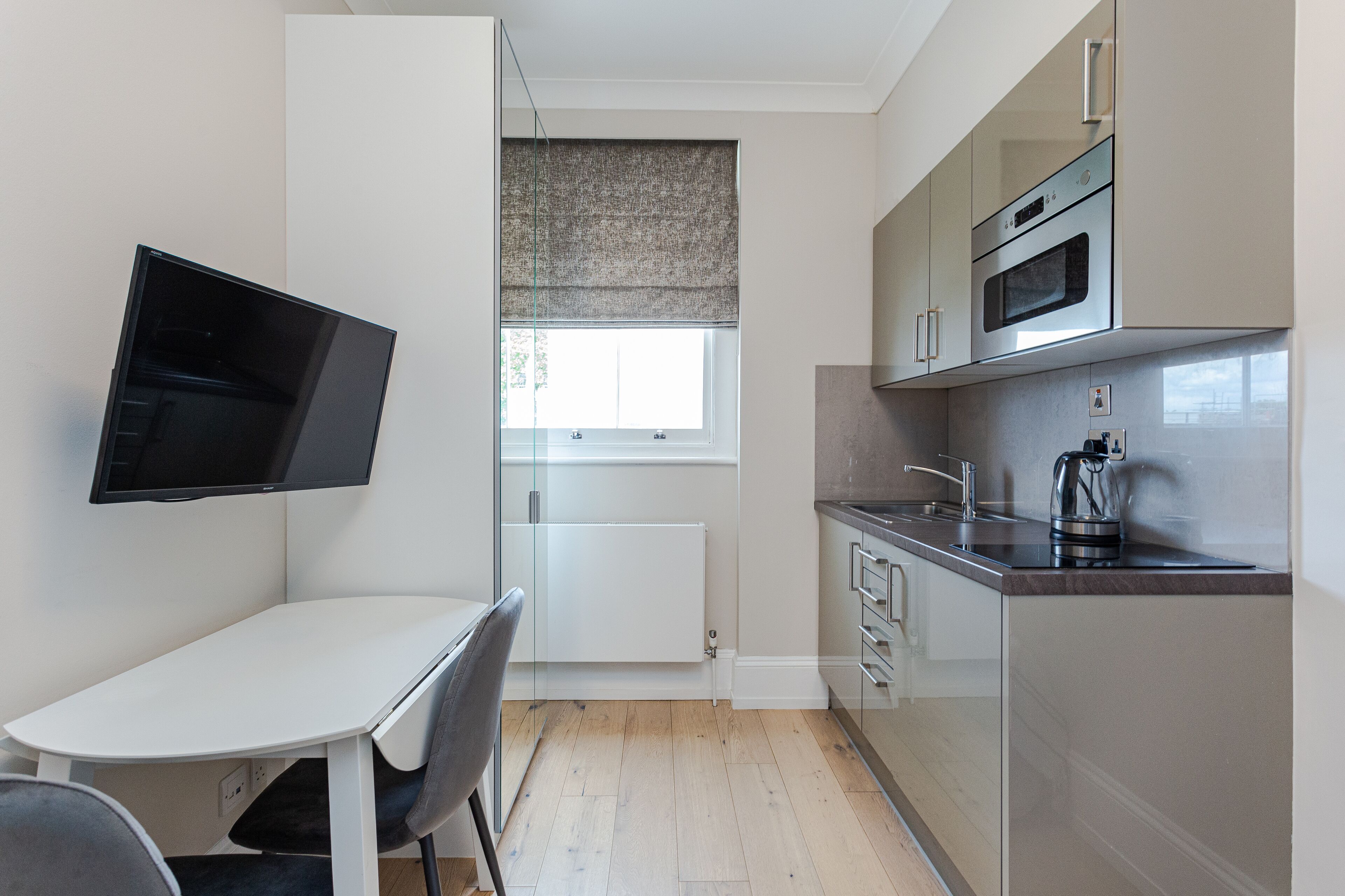 A modern kitchen with a built-in oven, microwave, and kettle. There is a dining table with chairs and a TV mounted on the wall.