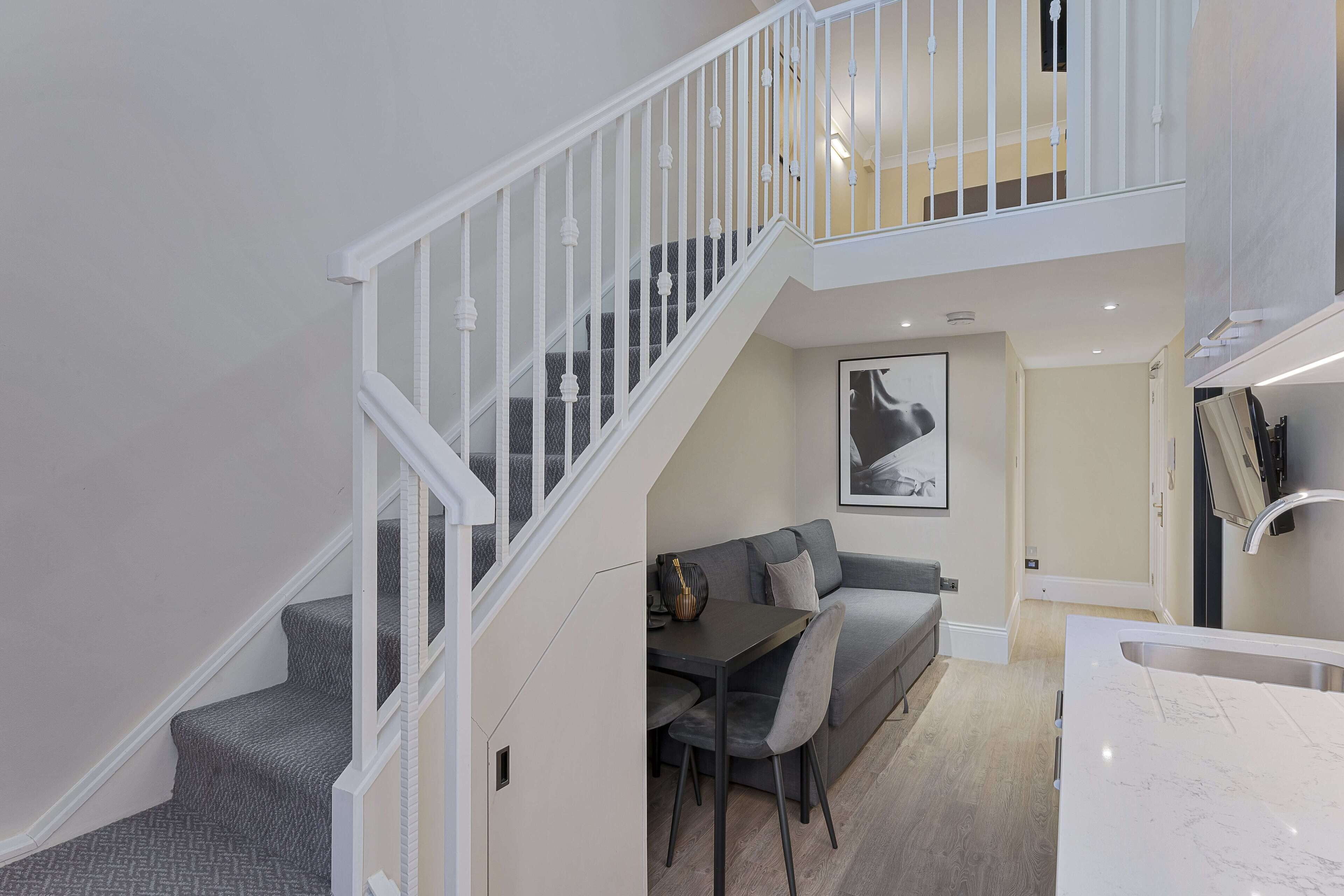 Ein modernes Interieur mit Treppe, Essbereich mit grauem Sofa und Tisch sowie Küche mit weißer Arbeitsplatte.