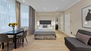 A modern hotel room with a bed, a sofa, a dining table with chairs, and a large window with curtains. - Cleveland Residences Kensington (London)