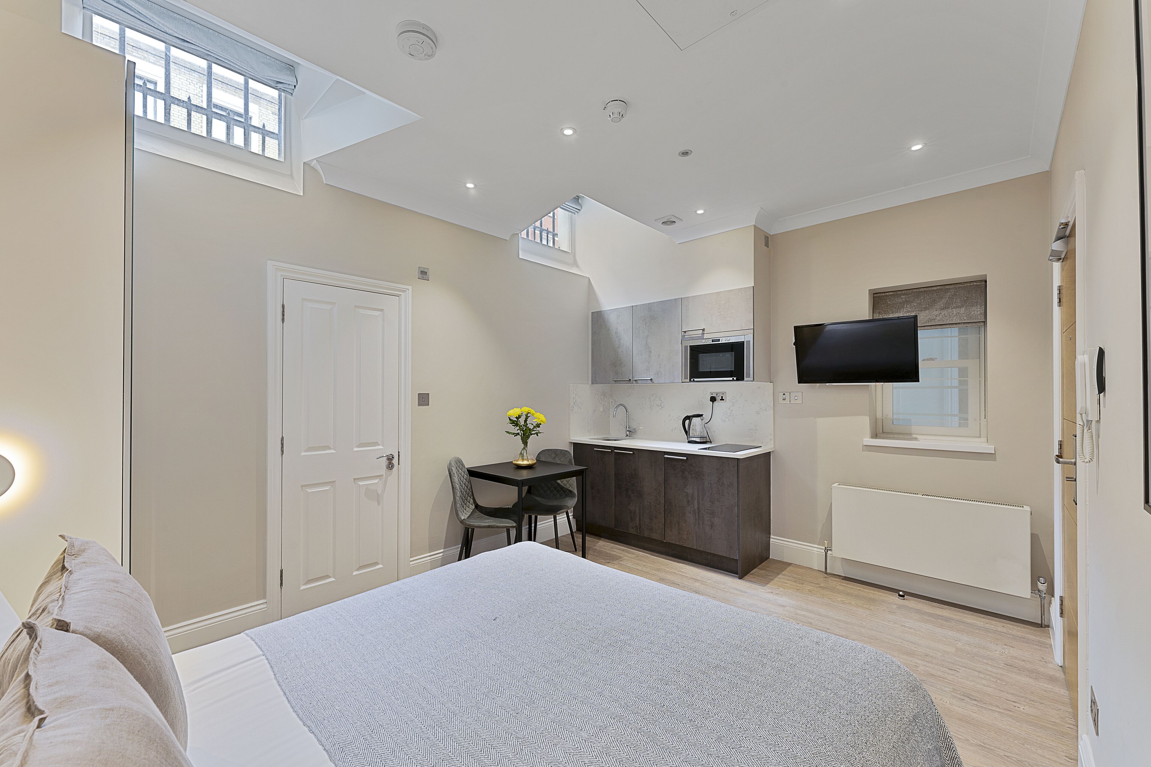 A modern bedroom with a large bed, a small dining table with chairs, a kitchenette, and a TV mounted on the wall.