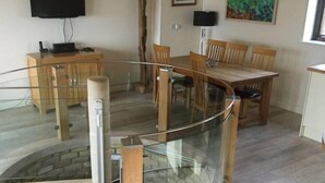 A dining area with a glass railing, wooden chairs, a table, and a TV mounted on the wall. - The Fish Cellars - Luxury Holiday Cottage (Portwrinkle)