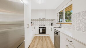 A compact kitchen with white cabinets, a built-in oven, and a window with a view of greenery. - 3 Haig Place - 100m from Kirra Beach with Wi-Fi included (Coolangatta)