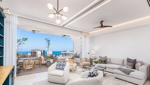 Una sala de estar moderna con un sofá, sillones y una mesa de centro. Se ve el océano y edificios al fondo.