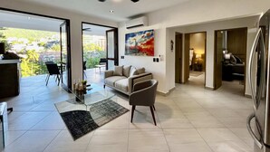 A modern living room with a glass coffee table, a grey armchair, and a view of a balcony with a cityscape.