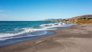 Una playa arenosa con olas, cielo despejado y colinas a lo lejos.