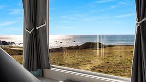 Una habitación con vista al océano a través de una ventana con cortinas grises recogidas.