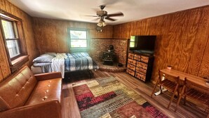 A cozy room with a bed, a sofa, a TV, a fireplace, and a ceiling fan. - Rustic Tabor Woods Cabin near Cacapon State Park w/Patio, Grill (Berkeley Springs)