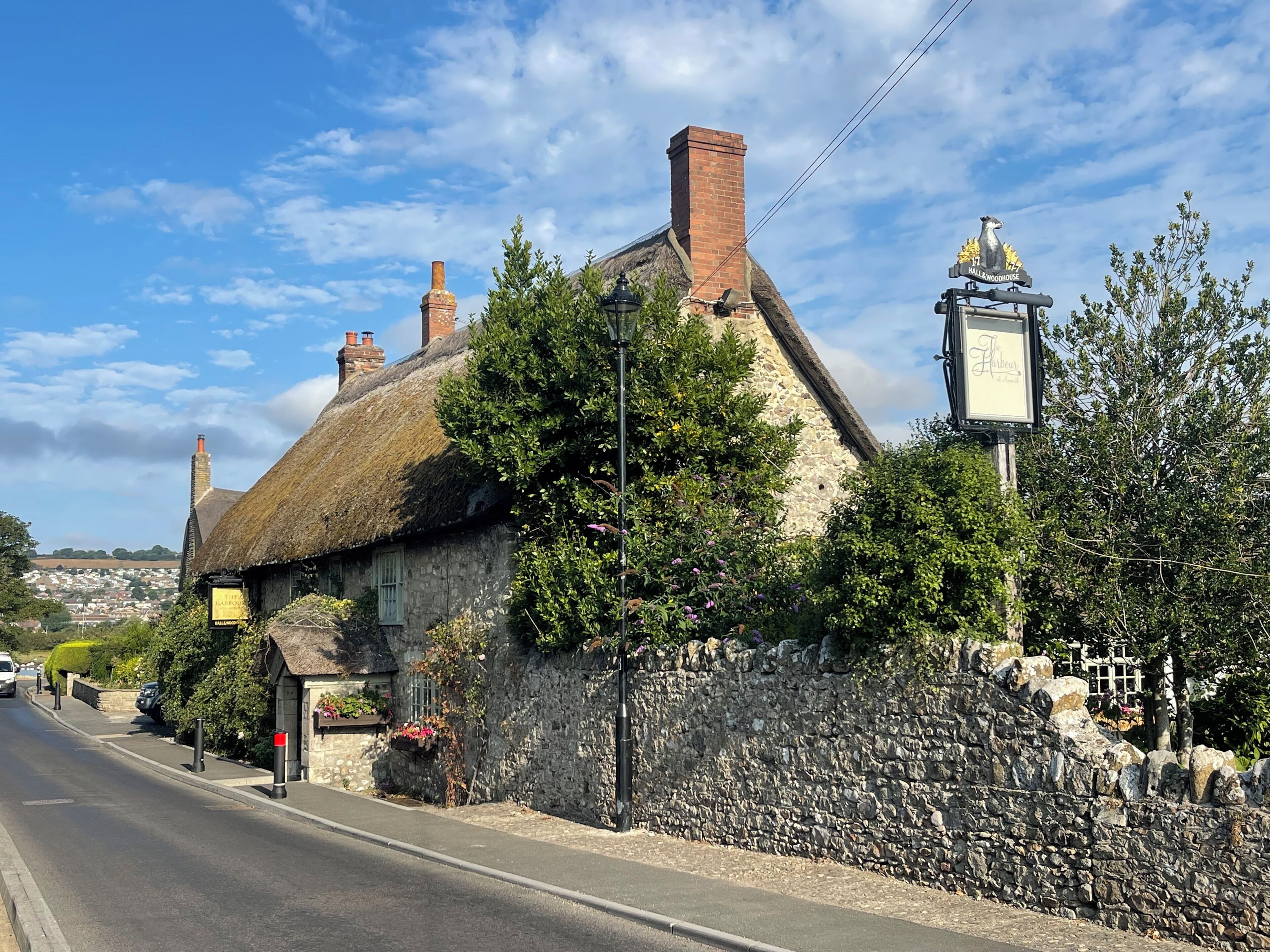 A traditional thatched-roof building with a stone wall, a street lamp, and a sign indicating a pub named 'Hallow Woodhouse.'