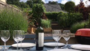 Mesa al aire libre con platos, copas de vino y cubiertos, una botella de vino y una parrilla de barbacoa al fondo.