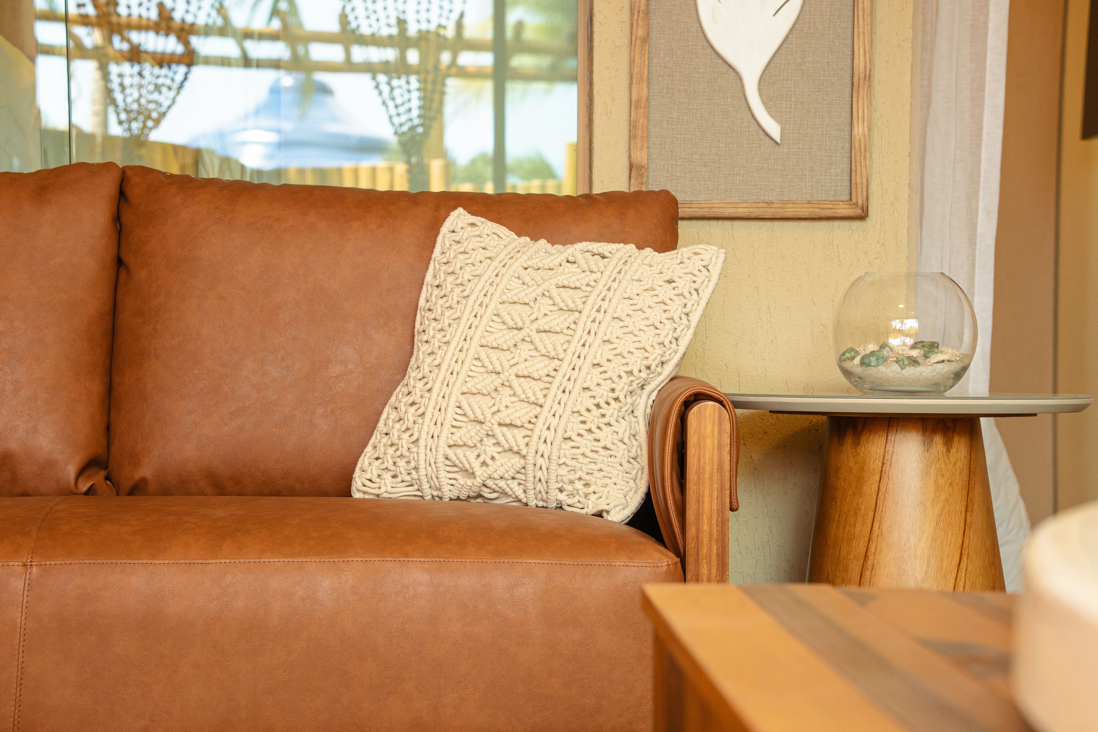 A brown leather sofa with a textured white pillow, a small wooden side table with a glass bowl, and a framed artwork on the wall.