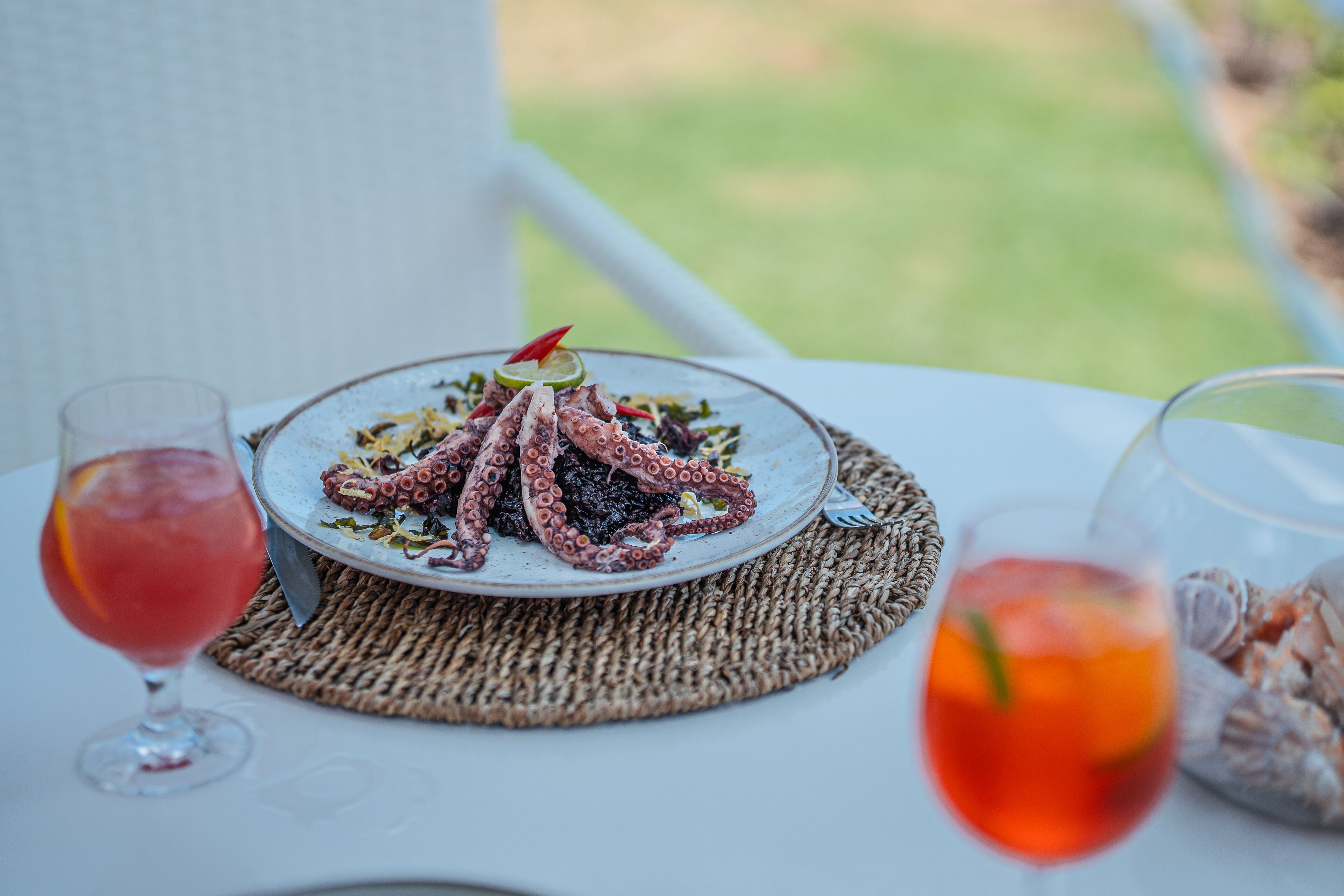 A table with a plate of octopus, two glasses of red drink, and a basket of seashells.