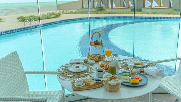 A dining table with breakfast items and a glass of orange juice by a pool with a view of the ocean. - Pousada Mansão da Costa (Maragogi)