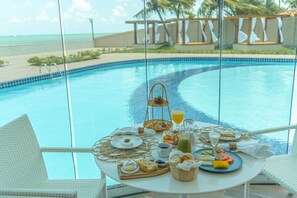 A dining table with breakfast items and a glass of orange juice by a pool with a view of the ocean. - Pousada Mansão da Costa (Maragogi)
