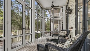 A covered porch with wicker furniture, a ceiling fan, and large windows offering a view of the surrounding trees.
