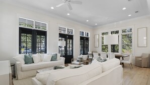 A spacious living room with a white sofa set, a dining area with a table and chairs, and large windows with black shutters. - Close to Parks with Private Pool and Bikes (Santa Rosa Beach)