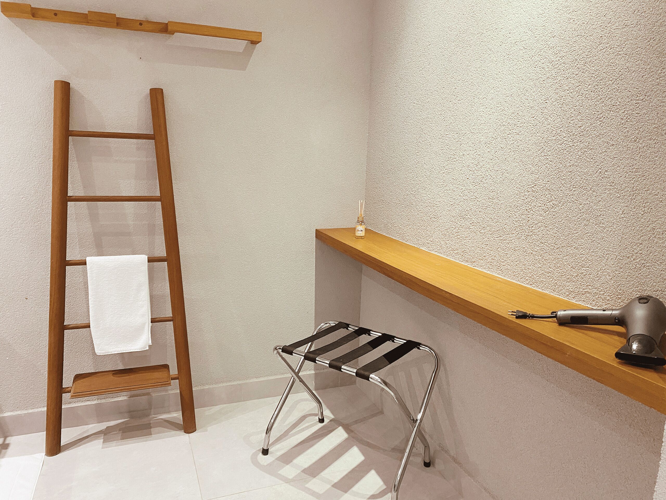 Un coin de pièce avec une échelle en bois, un petit tabouret, une étagère sur laquelle se trouve un sèche-cheveux, et une serviette accrochée à l’échelle.