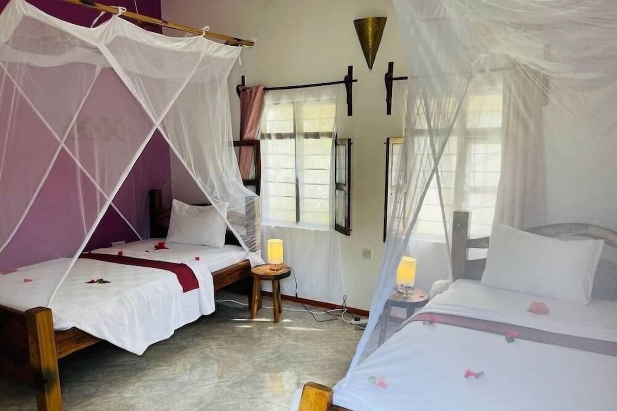 A room with two beds, each with a canopy, a small table with a lamp, and a window with curtains.