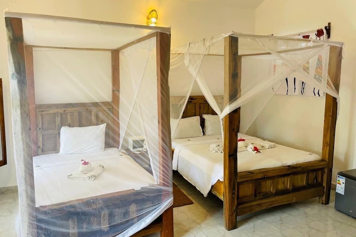 Two wooden four-poster beds with white linens and mosquito netting, a bedside table, and a small bedside lamp.