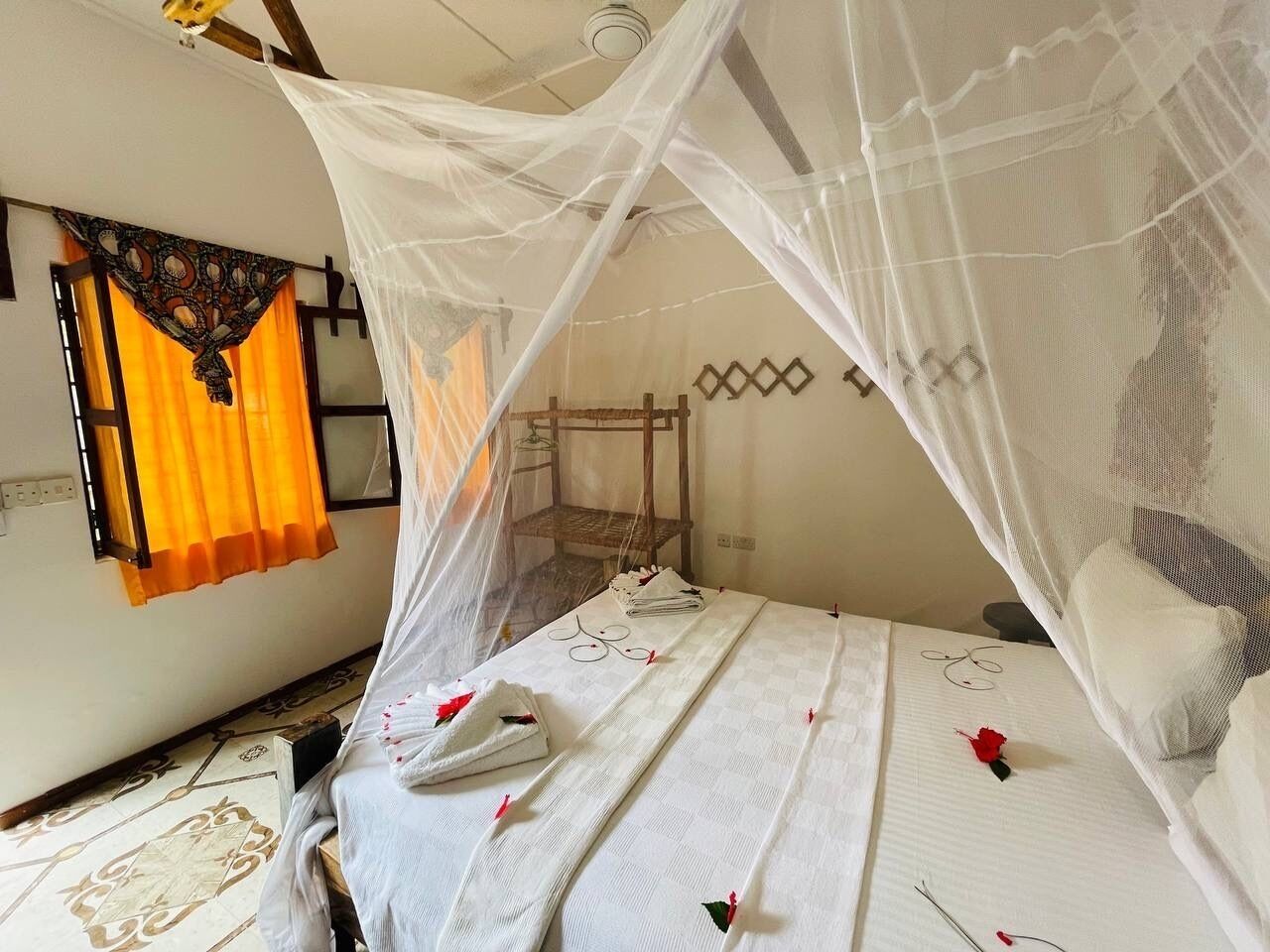 A room with a bed under a white canopy, a wooden chair, and a small table with a tray of towels and a red rose.