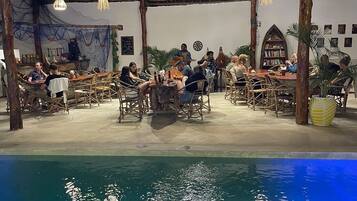An outdoor dining area with tables and chairs, a pool, and a thatched roof structure.