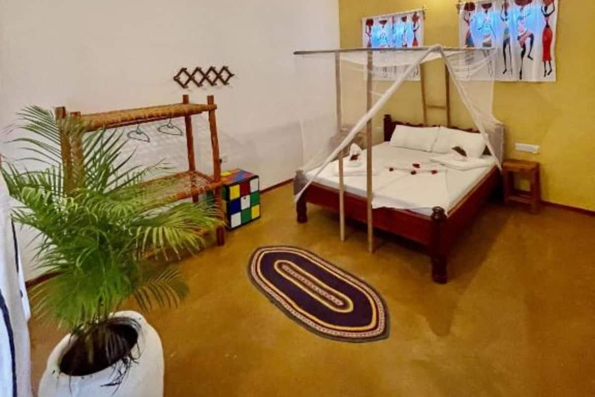 A room with a bed, a ceiling fan, a wall with artwork, a small table, and a potted plant.