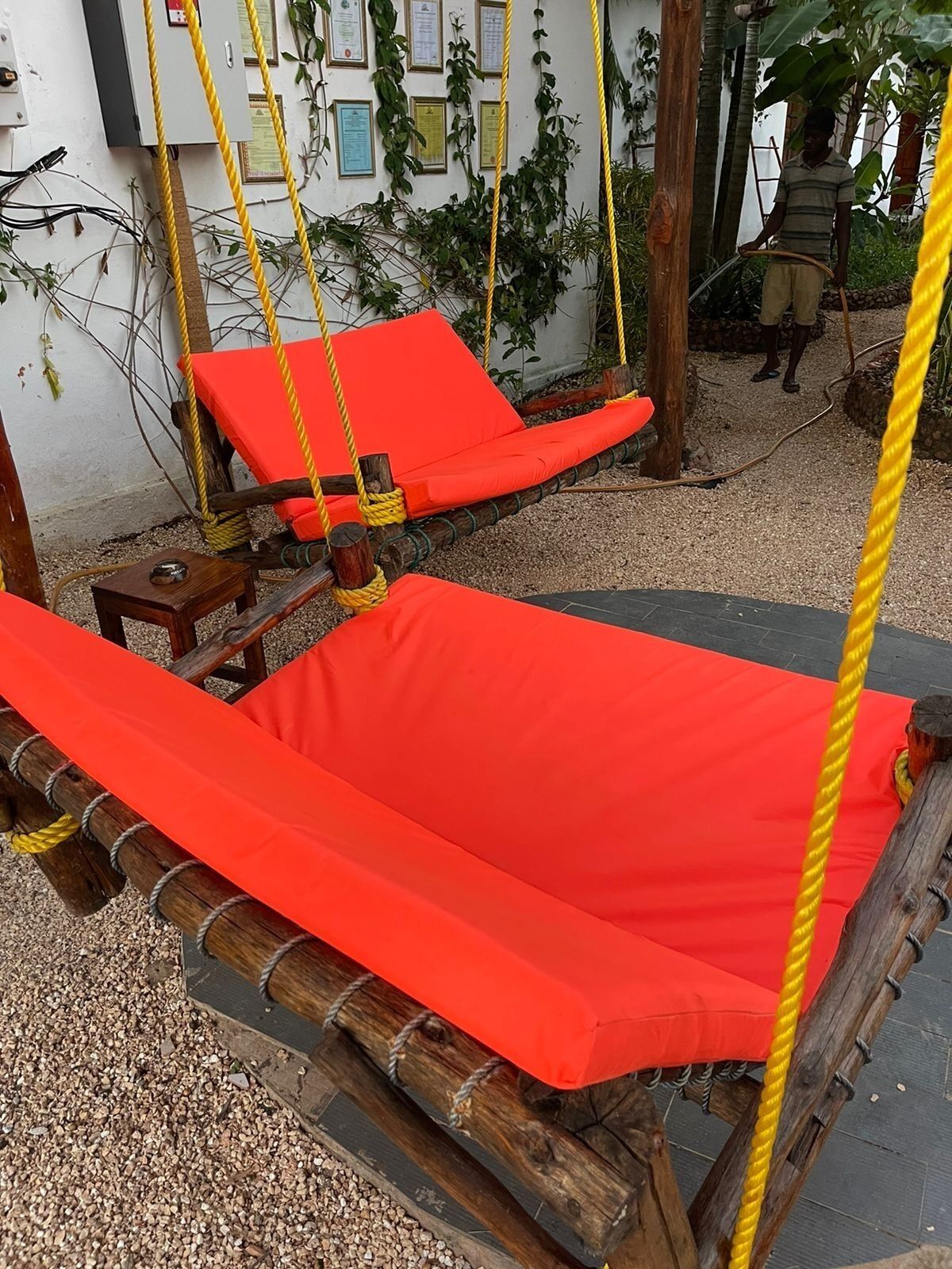 Two red cushioned hammocks suspended by yellow ropes, wooden footrests, and a wall with certificates and plants.