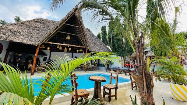 A pool area with a thatched-roof structure, wooden furniture, and tropical plants. - Babalao Bungalows (Nungwi)