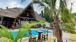 A pool area with a thatched-roof structure, wooden furniture, and tropical plants. - Babalao Bungalows (Nungwi)