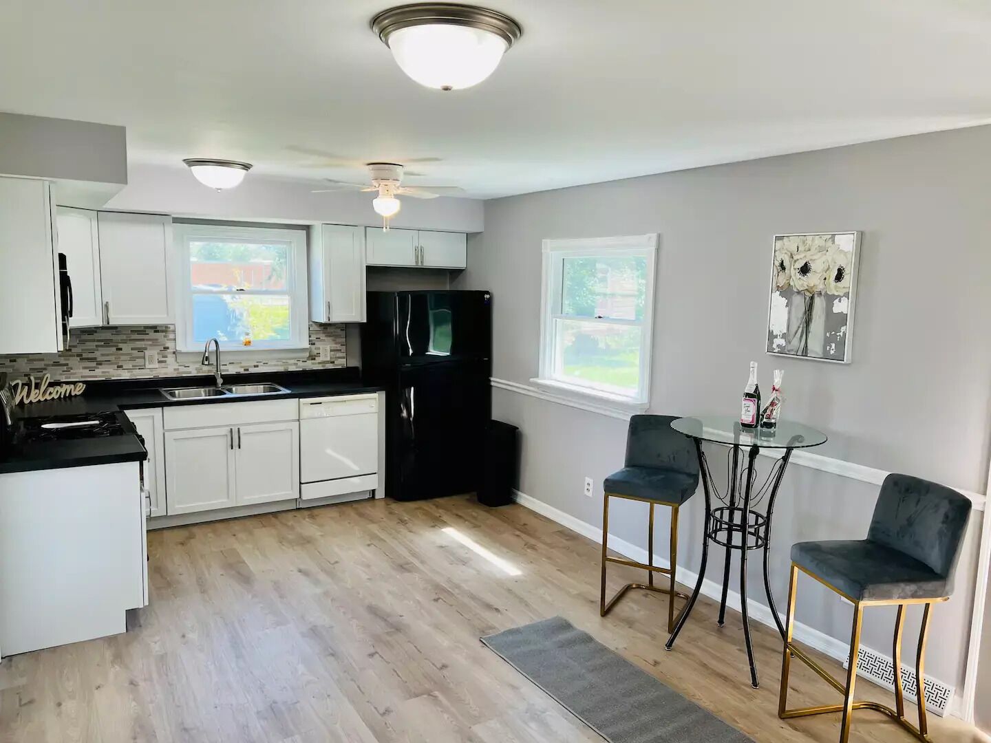 Een moderne keuken met witte kastjes, een zwarte koelkast, een plafondventilator en een eethoek met twee stoelen en een kleine tafel.