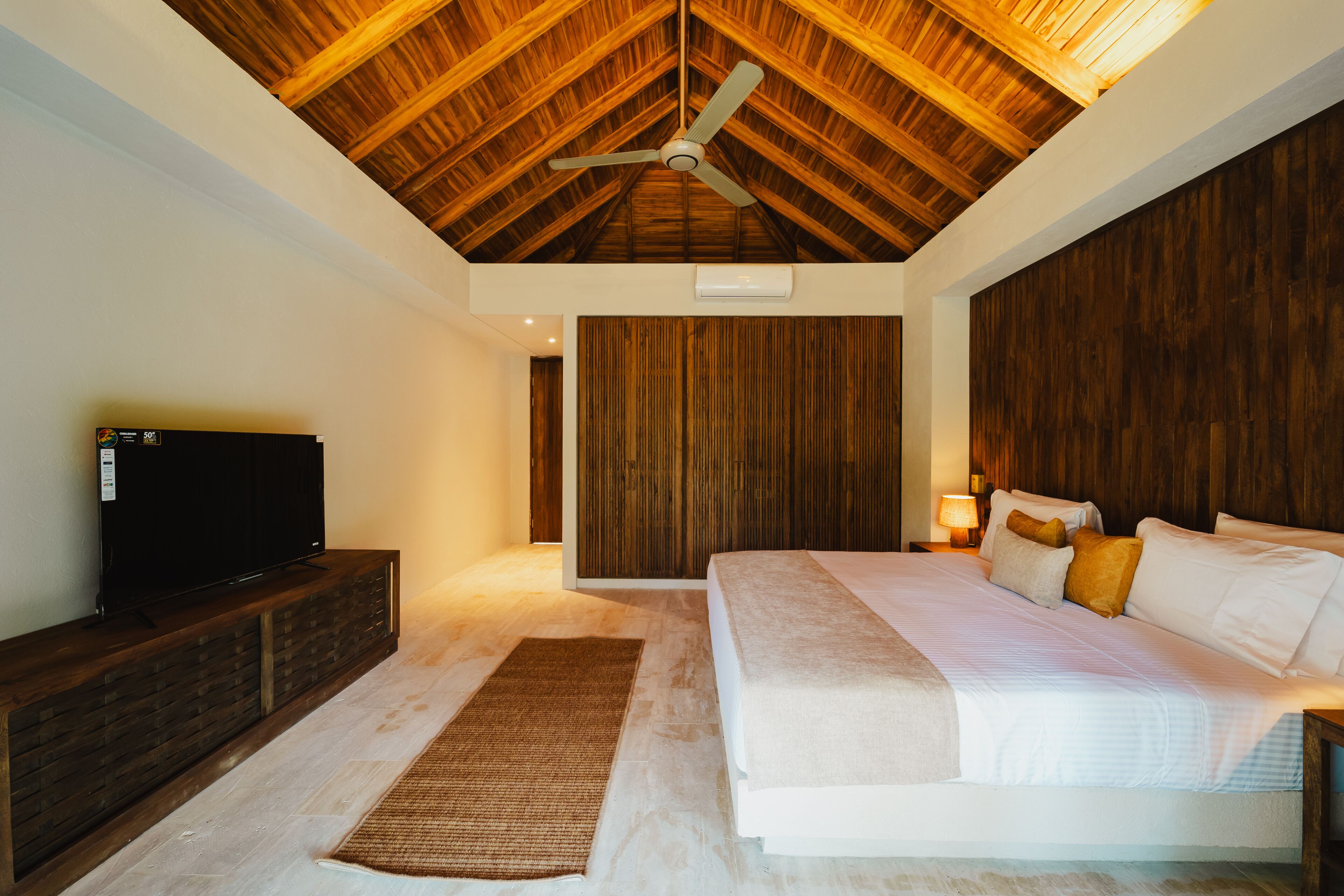 Un dormitorio con techo de madera, una cama con ropa blanca, un televisor sobre un mueble de madera y un panel de pared de madera.