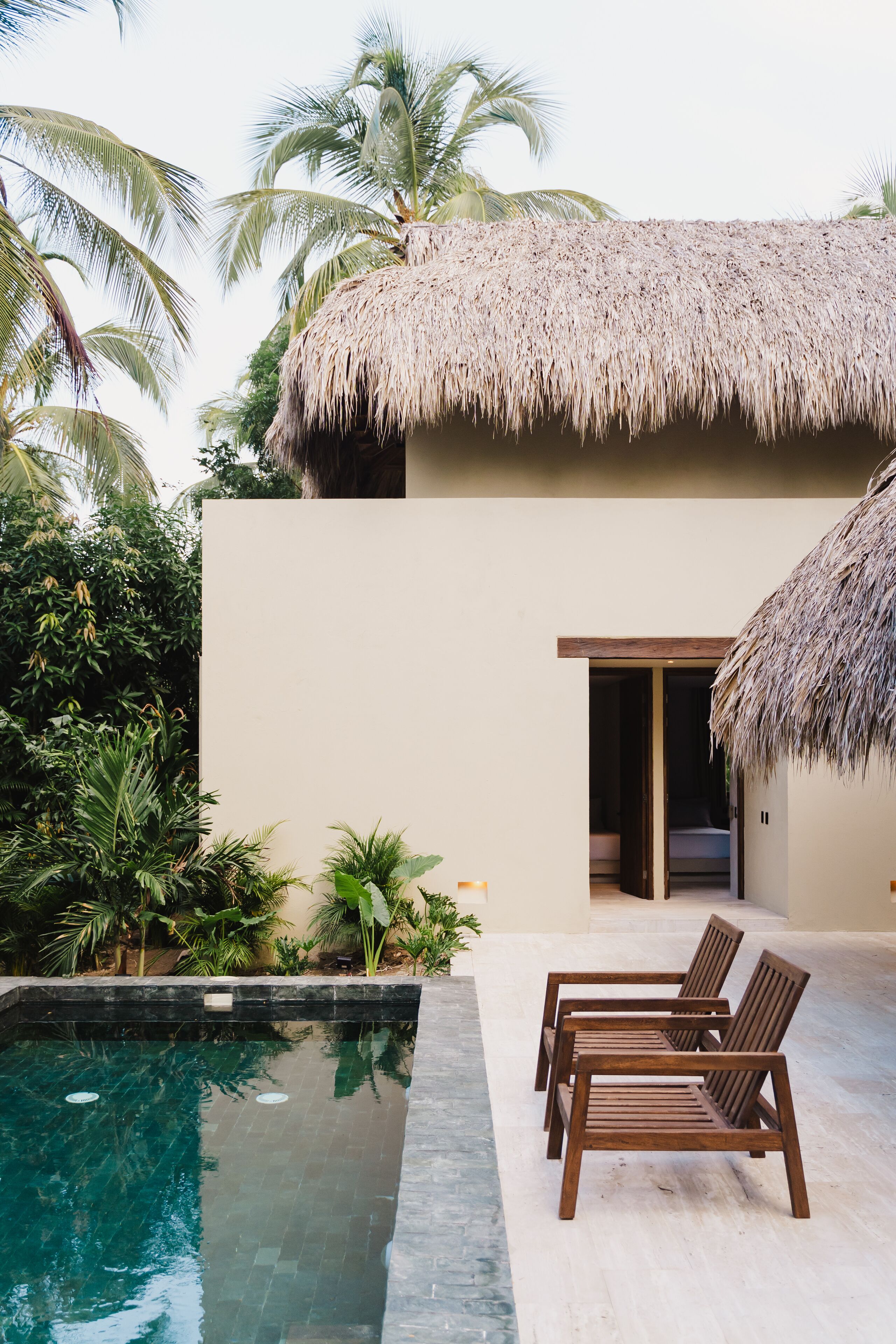 A pool area with a thatched roof structure, wooden lounge chairs, and lush tropical vegetation.