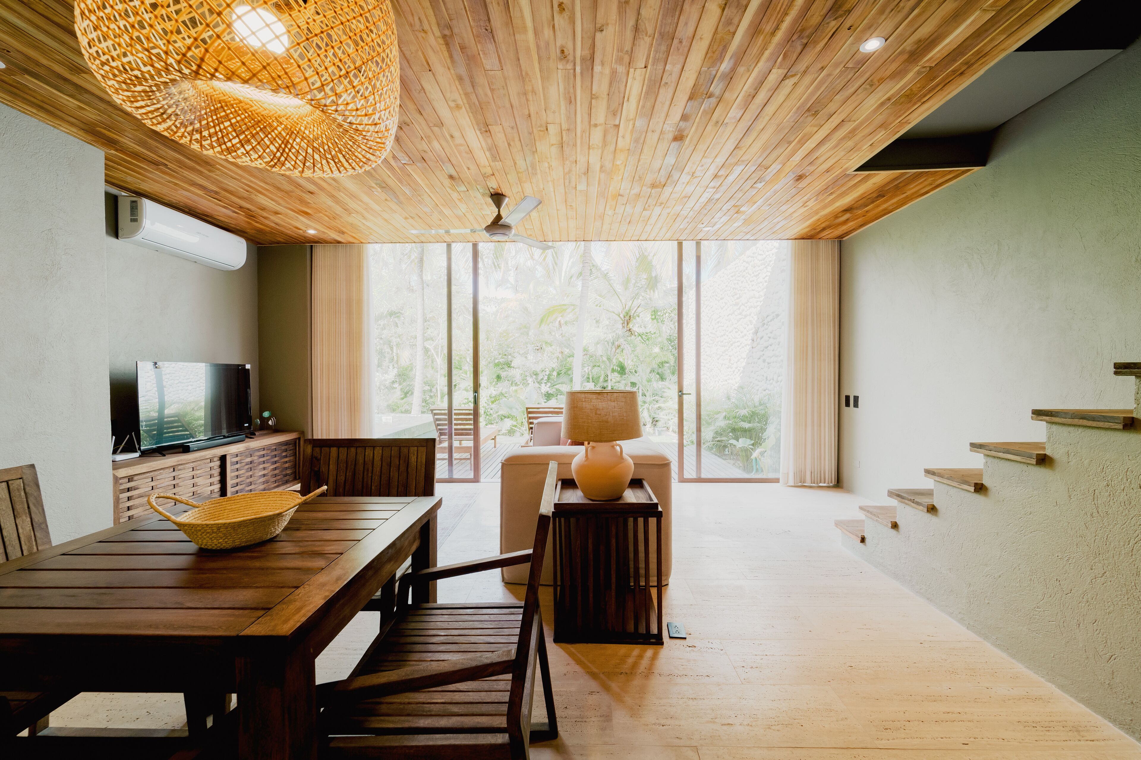 A modern living room with a wooden ceiling, a television, a dining table, and a large window offering a view of greenery.
