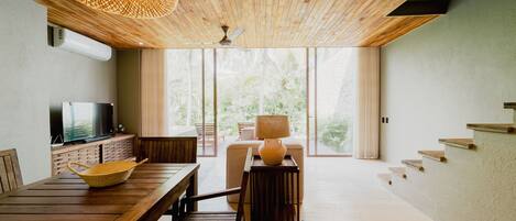 A modern living room with a wooden ceiling, a television, a dining table, and a large window offering a view of greenery.