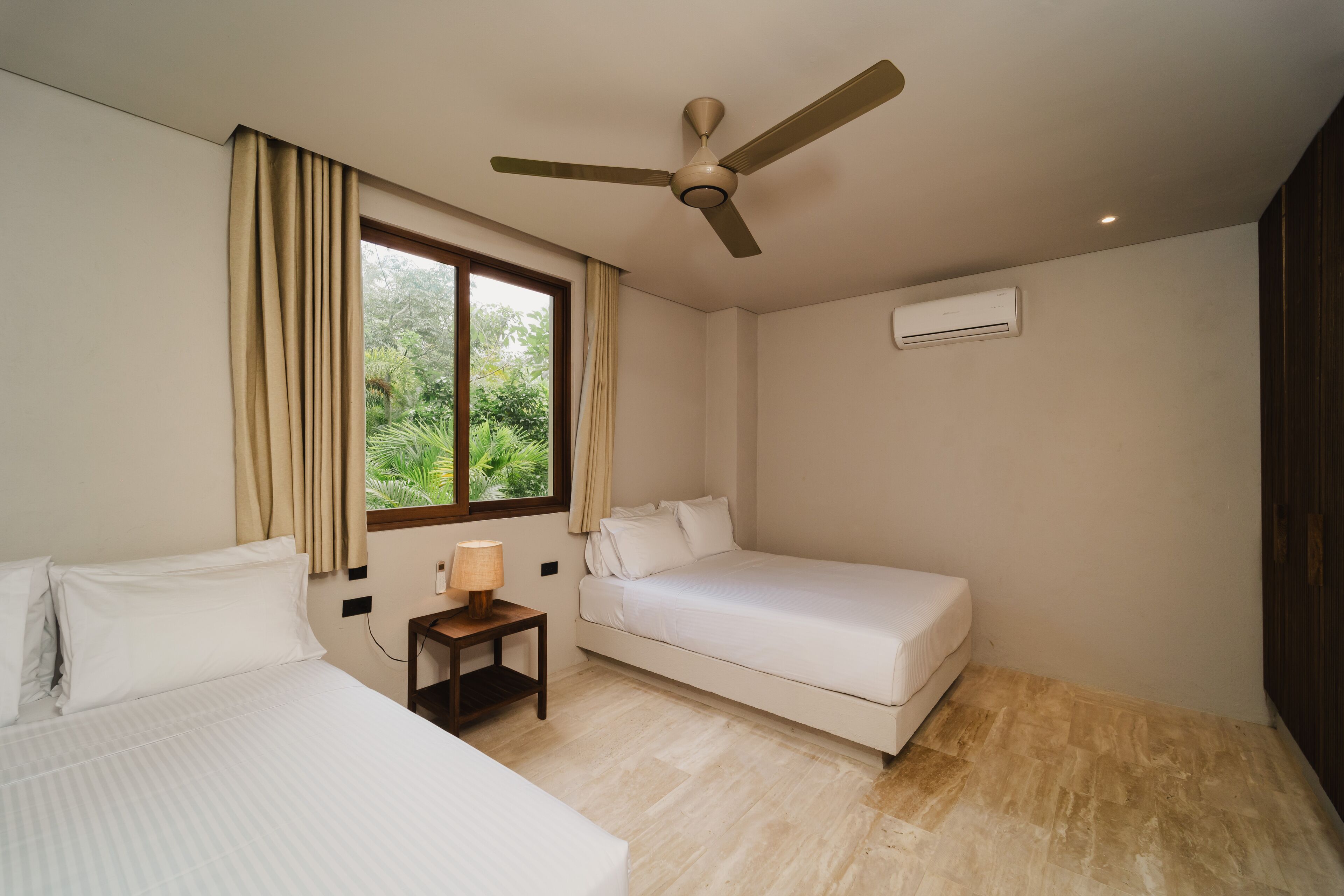 Ein Hotelzimmer mit einem Bett, einem Einzelbett, einem Nachttisch mit Lampe, einem Deckenventilator, einem Fenster mit Vorhängen und einer Klimaanlage.