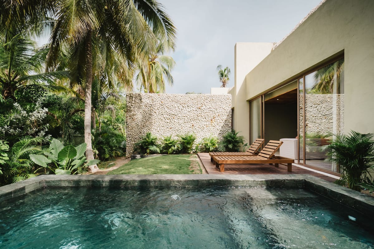A modern outdoor pool area with a stone wall, wooden lounge chairs, and lush tropical plants.