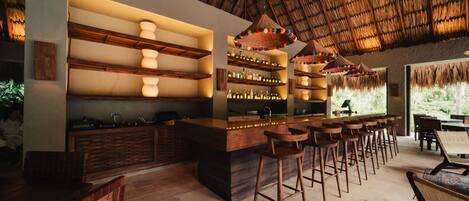 Zona de bar con estantes de madera, taburetes y techo con techo de paja.