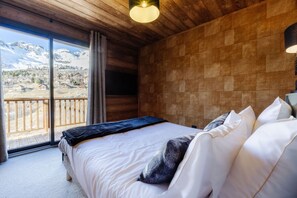 Une chambre avec un grand lit, un plafond en bois et une vue sur les montagnes, visible à travers une porte coulissante.