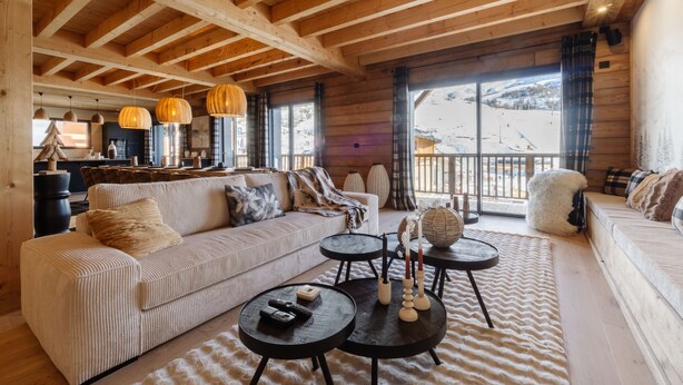 L’intérieur chaleureux d’un chalet en bois comprend un canapé, deux tables rondes et offre une vue sur un paysage enneigé grâce à de grandes fenêtres.